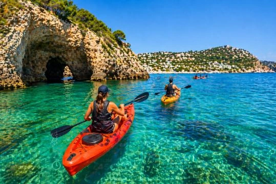 Personas haciendo kayak en aguas cristalinas frente a una cueva marina en la costa de Moraira (Alicante), rodeados de acantilados y paisaje mediterráneo.