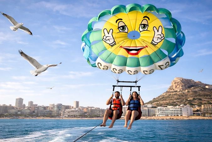 Dos turistas practicando paravelismo en Alicante, volando sobre el mar con un paracaídas colorido mientras varias gaviotas vuelan alrededor y el Castillo de Santa Bárbara se ve al fondo junto a la costa.