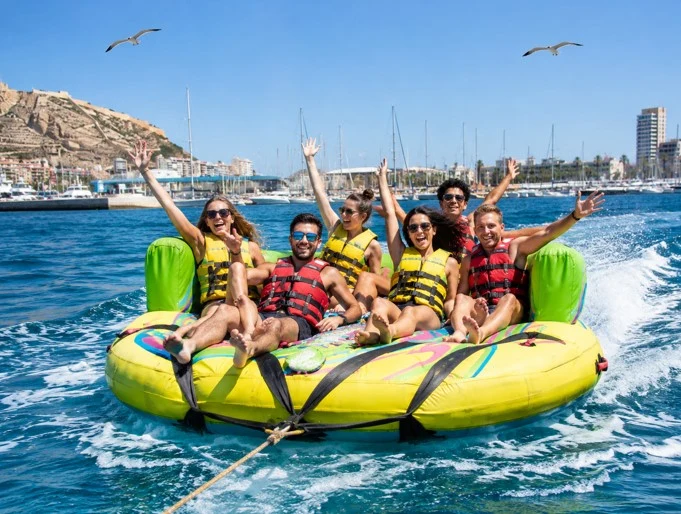 Grupo de turistas disfrutando de un sofá loco hinchable remolcado sobre el mar de Alicante, con chalecos salvavidas, gaviotas volando y el puerto al fondo.