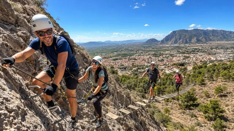 Grupo de excursionistas realizando la Vía Ferrata de Redován con casco y arnés, cruzando puentes colgantes sobre la montaña con vistas al paisaje de Alicante.