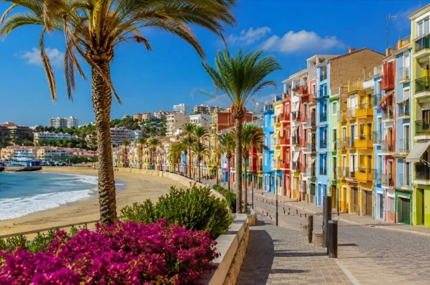 Casas de colores frente al mar en Villajoyosa, con paseo marítimo, palmeras y playa en un día soleado.